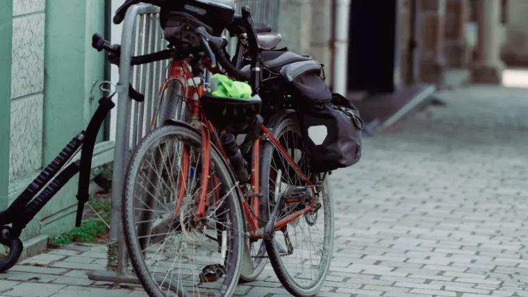 A red commuter bike with rear panniers and a helmet hanging from the handlebars, parked at a metal stand on a cobbled street