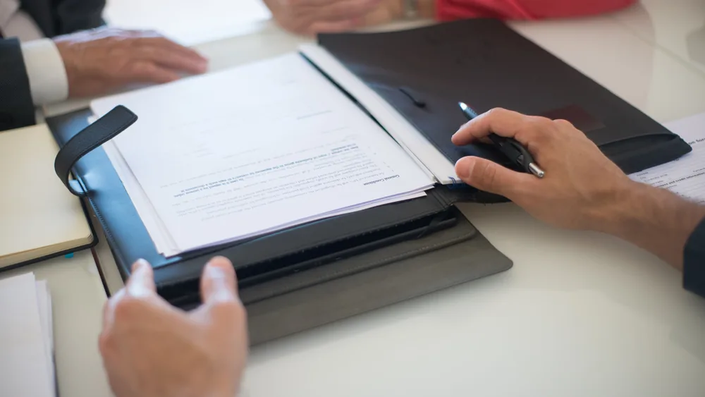 Hands resting on a table beside a folder of agreement papers and a pen