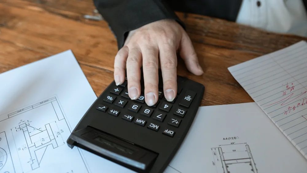 A hand using a calculator on a wooden desk covered with papers